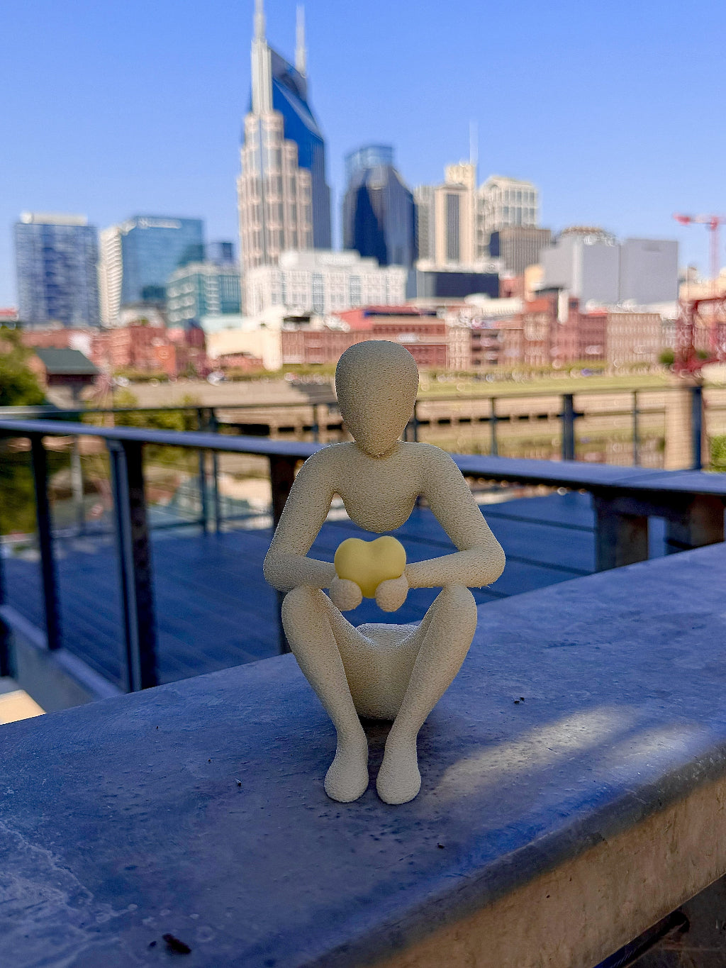 A 3D printed Nobody figure sits contemplatively on a railing overlooking the Nashville skyline, cradling a yellow 3D printed heart keychain in both hands.